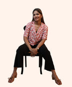 Symmetrical full-length front view of a woman posing on a chair in a peach pink daisy crop top with a notch collar by Ladymade.