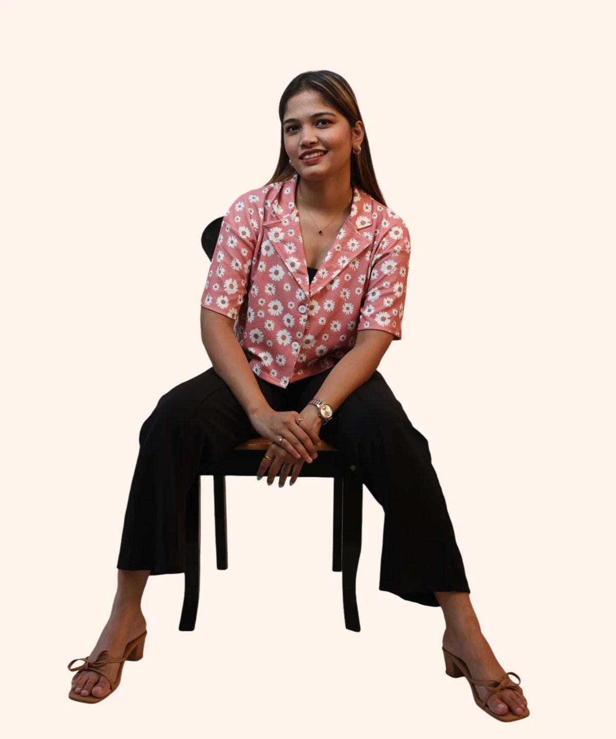 Symmetrical full-length front view of a woman posing on a chair in a peach pink daisy crop top with a notch collar by Ladymade.